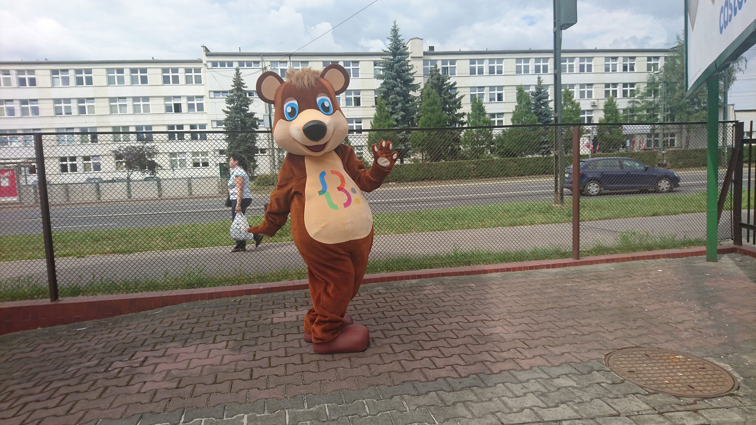 Na fotografii widnieje przebranie strój reklamowy Miś Tuliś pozujący do zdjęcia na jednej z krakowskich ulic. W tle przechodząca osoba, przejeżdżający samochód, budynek biurowy.